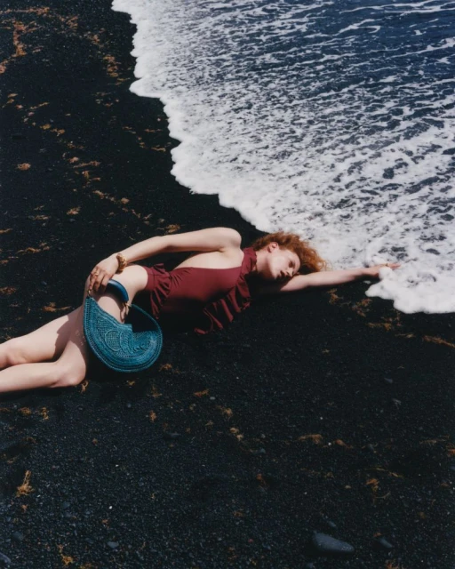 Woman in a swimsuit lying on a black sand beach with a blue crochet bag, with white foamy waves in the background.  