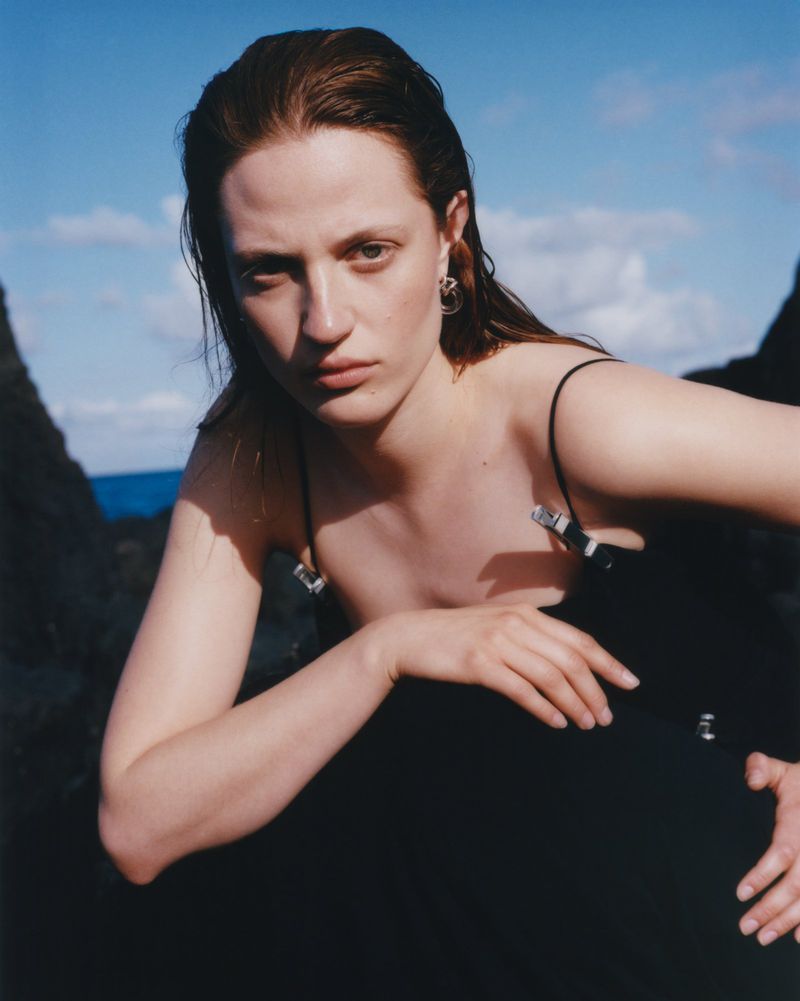 Portrait of an adult female model with brown hair and wearing a black dress, posing outdoors with a blue sky and rock formations.