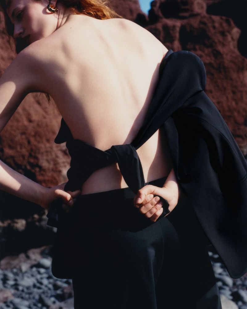 Back view of a redhead woman in black pants and top, standing outdoors with red rocks and blue sky in the background. Fashion shot.