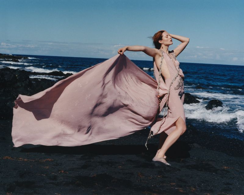 A woman poses with a flowing pink fabric on a black sand beach with ocean waves and a clear blue sky. She has red hair.