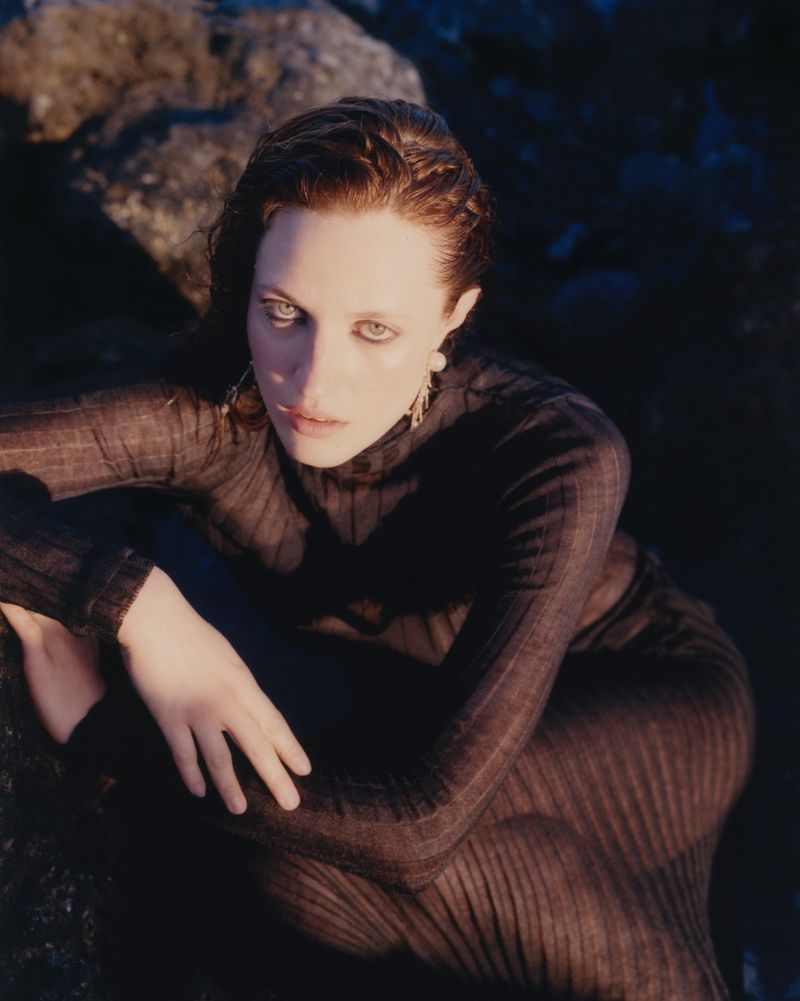 Eye-level shot of an adult woman with fair skin and wet hair wearing a dark, sheer dress sitting on dark rocks in a moody outdoor setting.