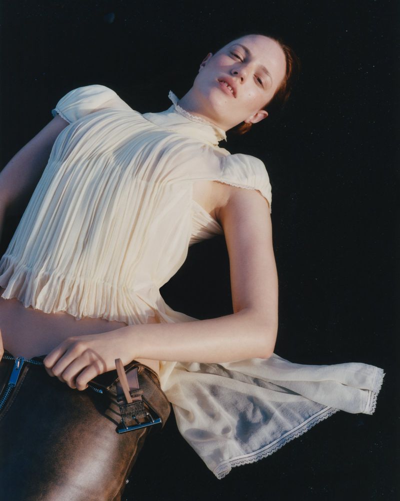 A woman with red hair poses in a pale top and brown pants against a dark background in an eye-level shot focusing on fashion.