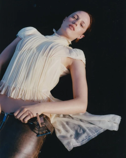A woman with red hair poses in a pale top and brown pants against a dark background in an eye-level shot focusing on fashion.