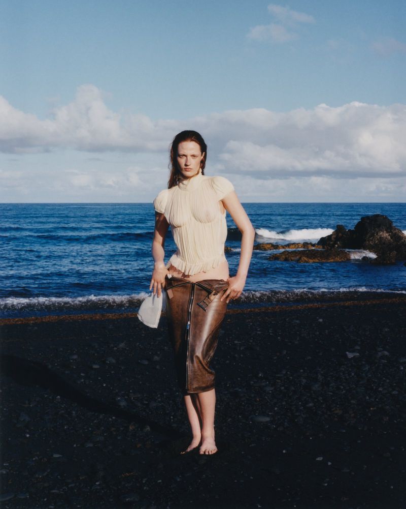 A bare-footed adult female fashion model poses on a black sand beach, wearing a light-colored corset and brown PVC capri pants, holding a bag.