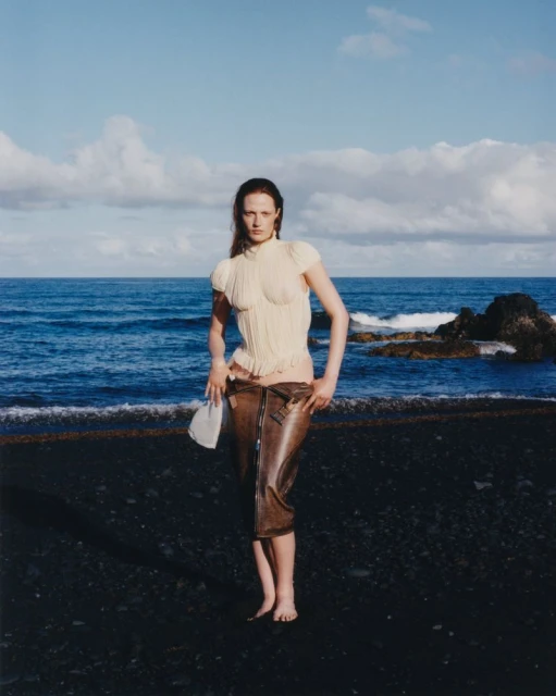A bare-footed adult female fashion model poses on a black sand beach, wearing a light-colored corset and brown PVC capri pants, holding a bag.