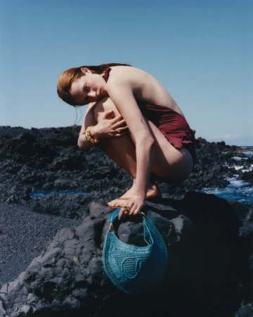 Model Julia Banas in a burgundy swimsuit crouching on dark coastal rocks holding a teal crochet bag under a clear sky.