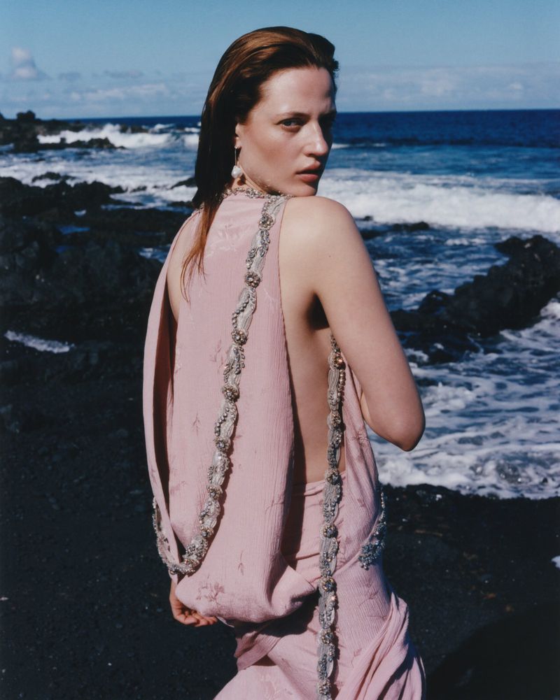 Woman in pink dress poses on black sand beach with ocean waves and blue sky behind her, looking over her shoulder at the camera.