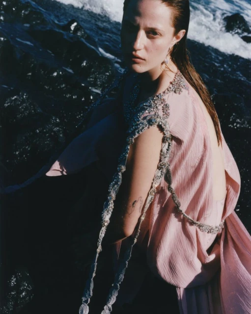 A young woman with wet hair poses in a flowing pink gown near the ocean, gazing directly at the viewer with a gentle expression.