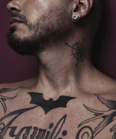 Close-up of a light-skinned adult male with tattoos on his chest and neck, a silver earring, and a short beard, against a plain background.