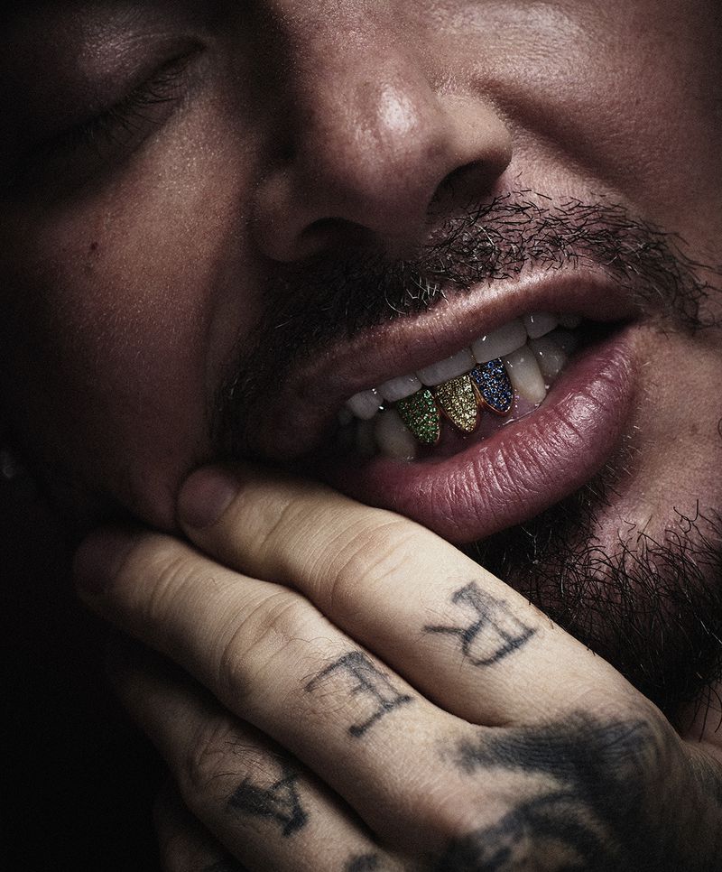 Close-up of a man's face with multi-colored diamond grillz on his teeth, and hand with tattoos covering his mouth, indoor setting.
