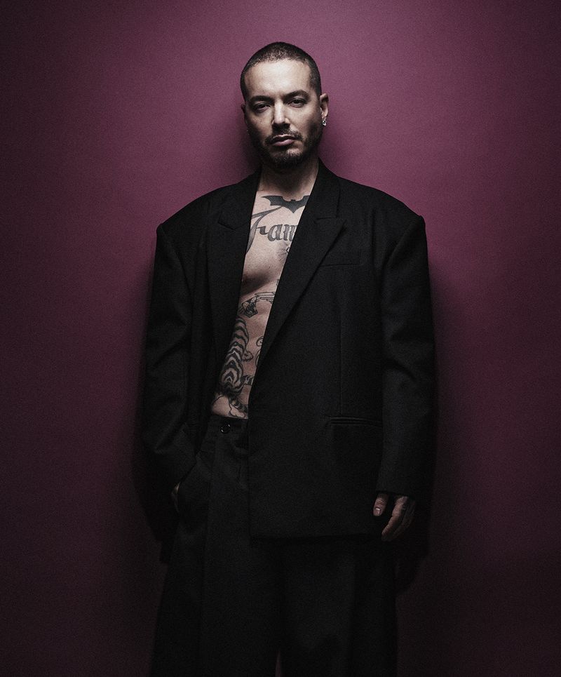 Portrait of a tattooed adult man in a black suit with an open jacket, posing against a solid dark pink backdrop in a studio setting.