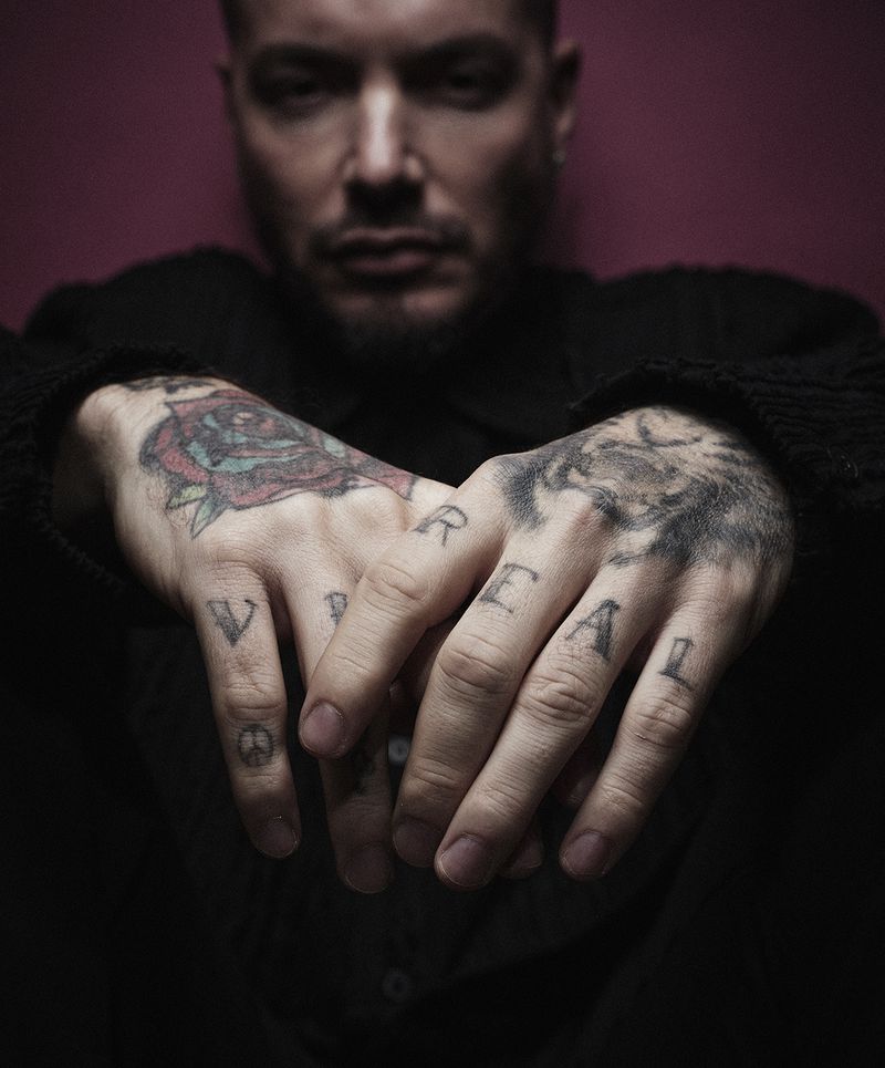 Adult male's hands with tattoo art and the word REAL on the fingers. He wears a black shirt and is in a dimly lit indoor setting.