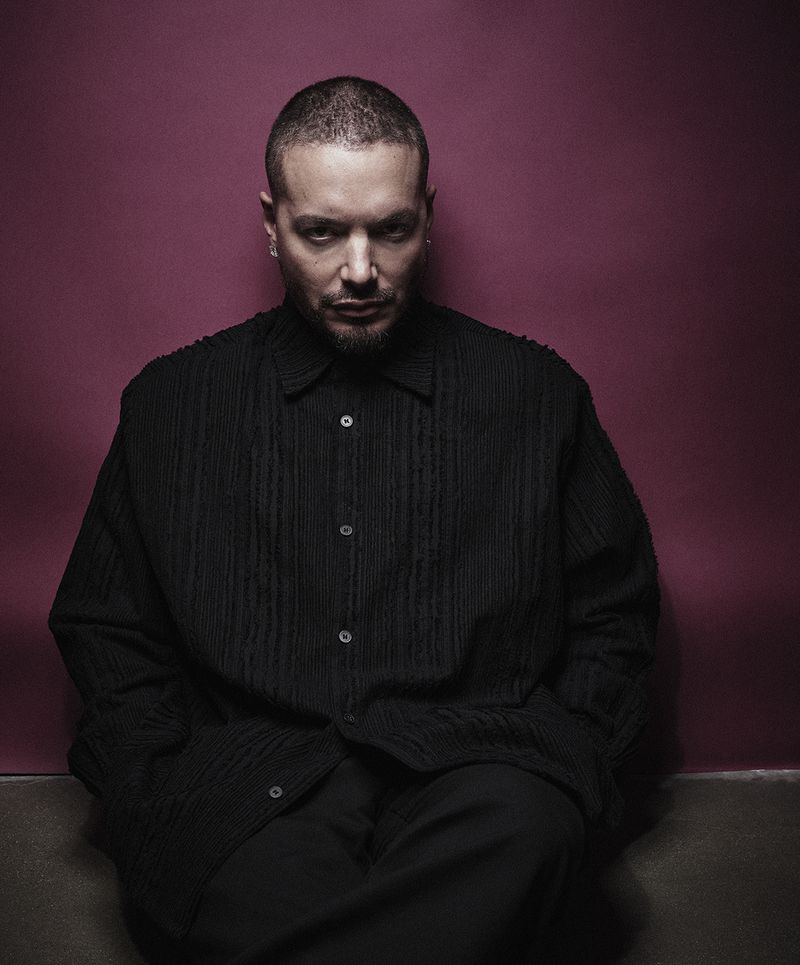 Portrait of an adult male wearing a black button-down shirt and pants, set against a pink and grey background, gazing at the camera.