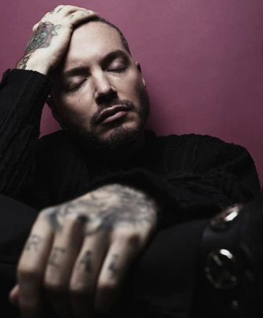 Close-up portrait of adult male with tattoos, eyes closed, wearing black top against magenta background in a dimly lit studio setting.