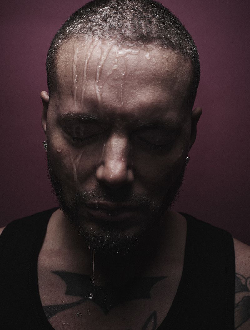 Close-up of a man with water dripping down his face, a tattoo on his chest, and a solid reddish-purple background creating a somber mood.