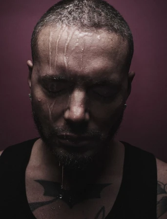 Close-up of a man with water dripping down his face, a tattoo on his chest, and a solid reddish-purple background creating a somber mood.