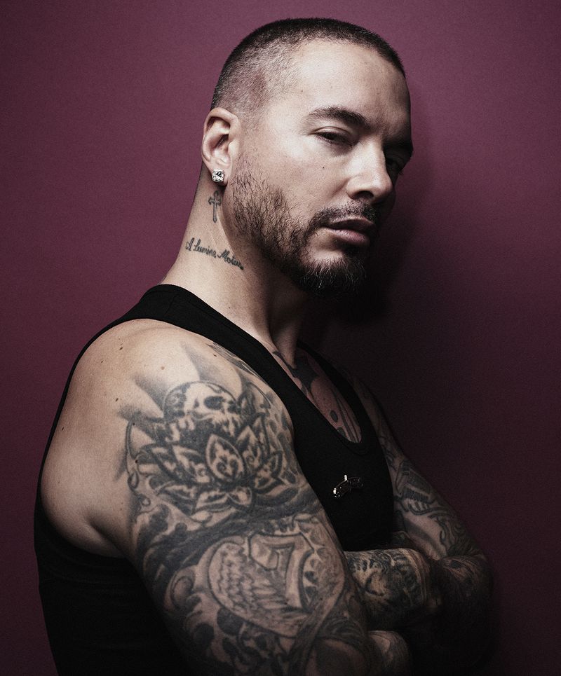 Portrait of an adult male with a buzz cut, tattoos, and arms crossed against a maroon background in a photo studio environment.