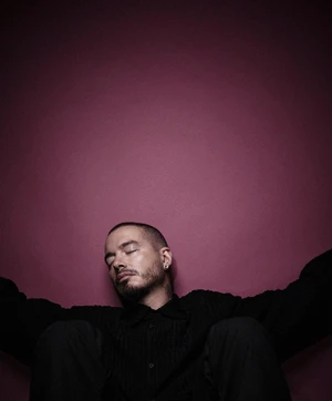 Portrait of musician J Balvin leaning back with eyes closed in a black shirt against a solid deep magenta backdrop.