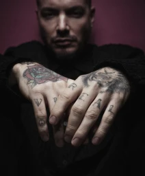Musician J Balvin holding his tattooed hands forward displaying knuckle tattoos against a dark red backdrop.
