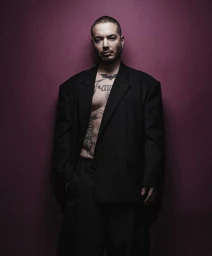 J Balvin wearing a loose black blazer over bare tattooed chest against a dark plum wall