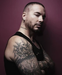 Close-up portrait of J Balvin with tattoos and a shaved head against a solid dark red background