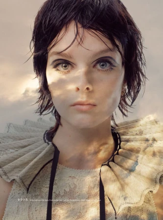 Model Aylah Peterson with short wet-look dark hair wearing a pleated lace ruff collar against a soft cloudy sky backdrop.