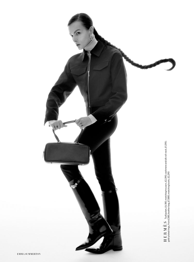 Aylah Peterson in a black zip jacket, trousers, and tall black riding boots holding a leather bag against a white studio background.