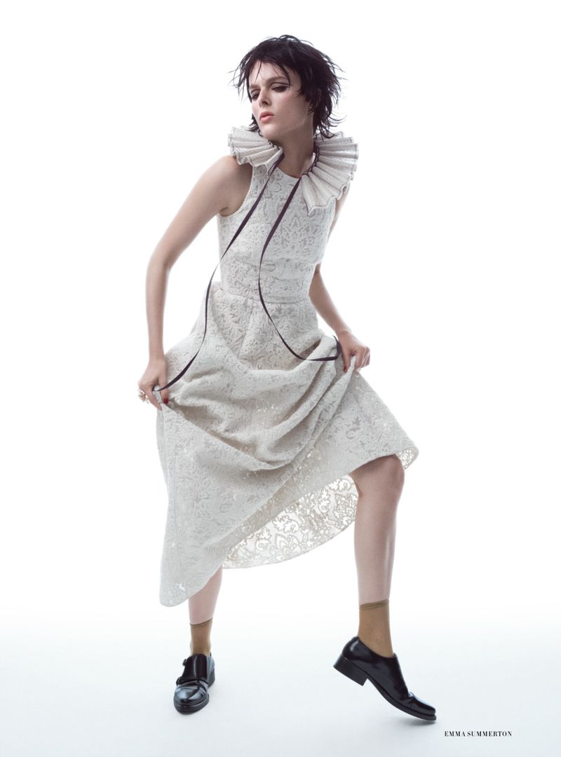 Editorial fashion shot of a woman in an off-white dress with a ruffled collar, standing in a white studio with one foot forward.