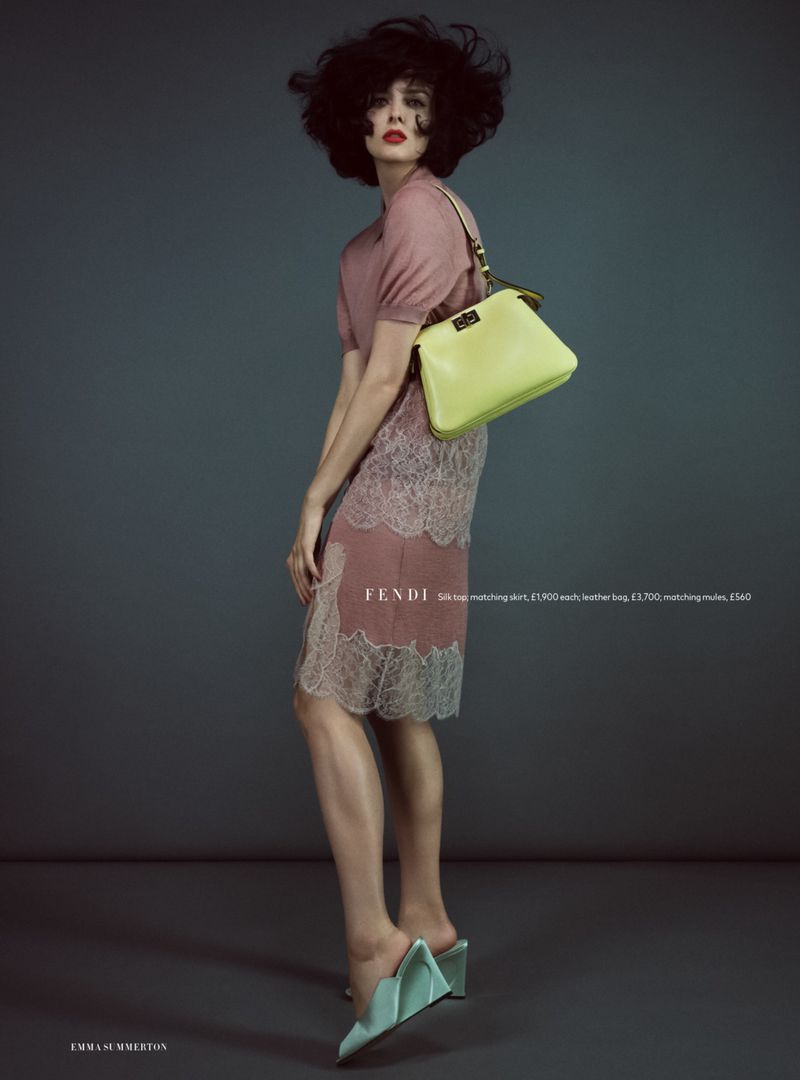 A woman poses in a fashion editorial shot, wearing a pink dress with lace, mint green shoes, and a lemon-yellow handbag. Studio setting.