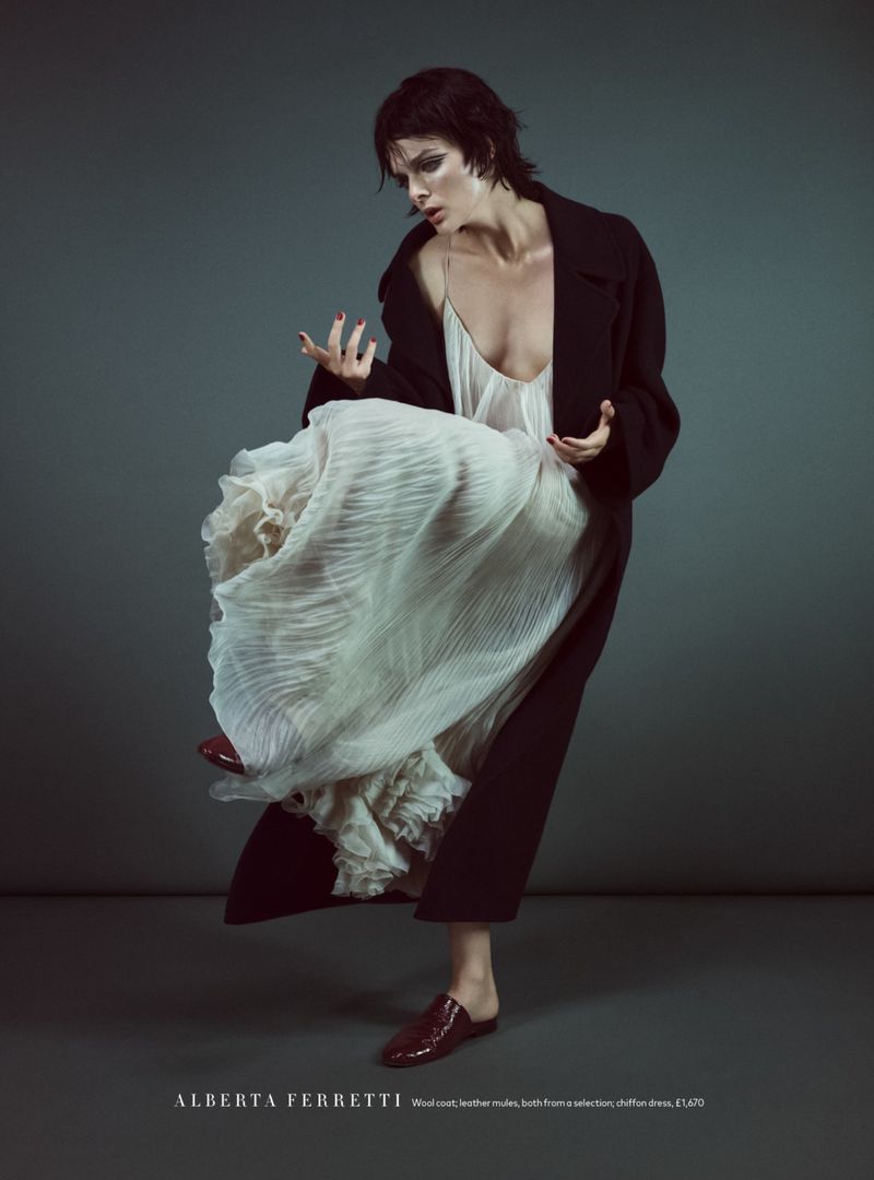 Model Aylah Peterson wearing a flowing white pleated dress, black overcoat, and dark red mules against a grey studio backdrop.