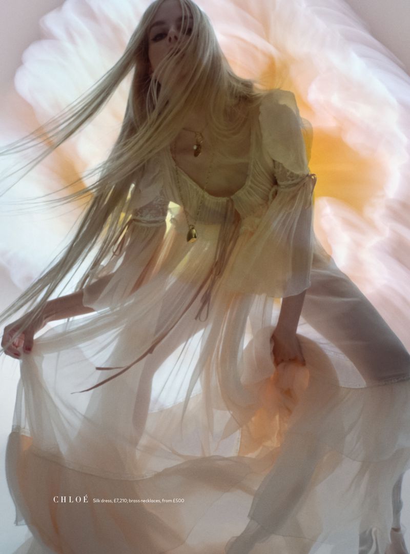 A fashionable adult female with long blonde hair wearing a sheer dress poses in front of a blurred floral background, fashion shot.