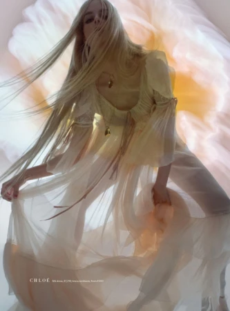 A fashionable adult female with long blonde hair wearing a sheer dress poses in front of a blurred floral background, fashion shot.