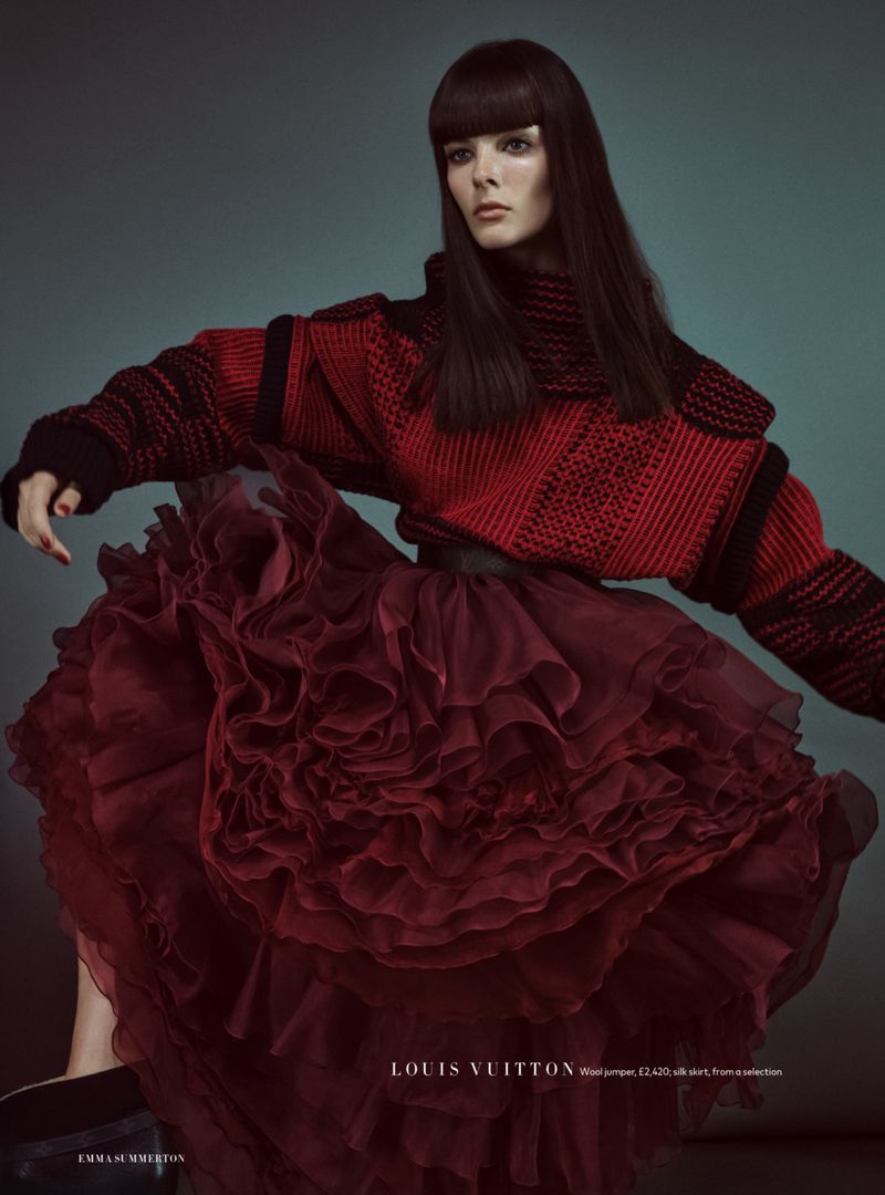 Fashion shot of a woman in a red sweater and ruffled skirt posing in front of a teal backdrop, showcasing dark hair and makeup.