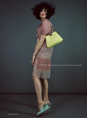 Model Aylah Peterson posing in a pink lace dress, voluminous black hair, and a lime green handbag against a grey backdrop.