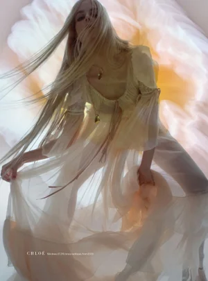 Blonde model in a sheer cream silk dress with flowing hair posing in front of a giant soft pink floral projection.