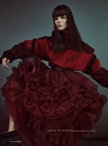 Model wearing a chunky red knit sweater and a voluminous ruffled burgundy skirt against a muted grey-green backdrop.