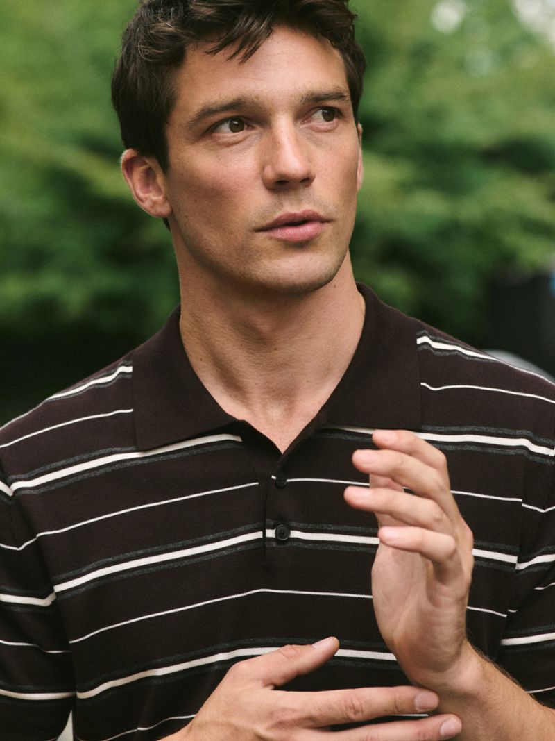 Portrait of a light-skinned adult male with dark hair wearing a striped polo shirt outside with a blurry green leafy background.