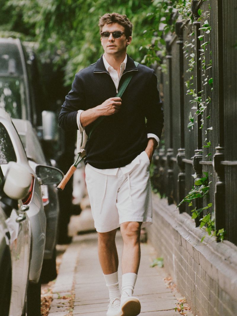 An adult male walks outdoors wearing a dark blue sweater, white shorts, socks, and sneakers, carrying a tennis racket on his shoulder.
