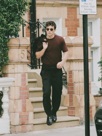 A confident man descends outdoor steps of brick townhouse, wearing a maroon shirt, black trousers, black jacket over shoulder in daylight.