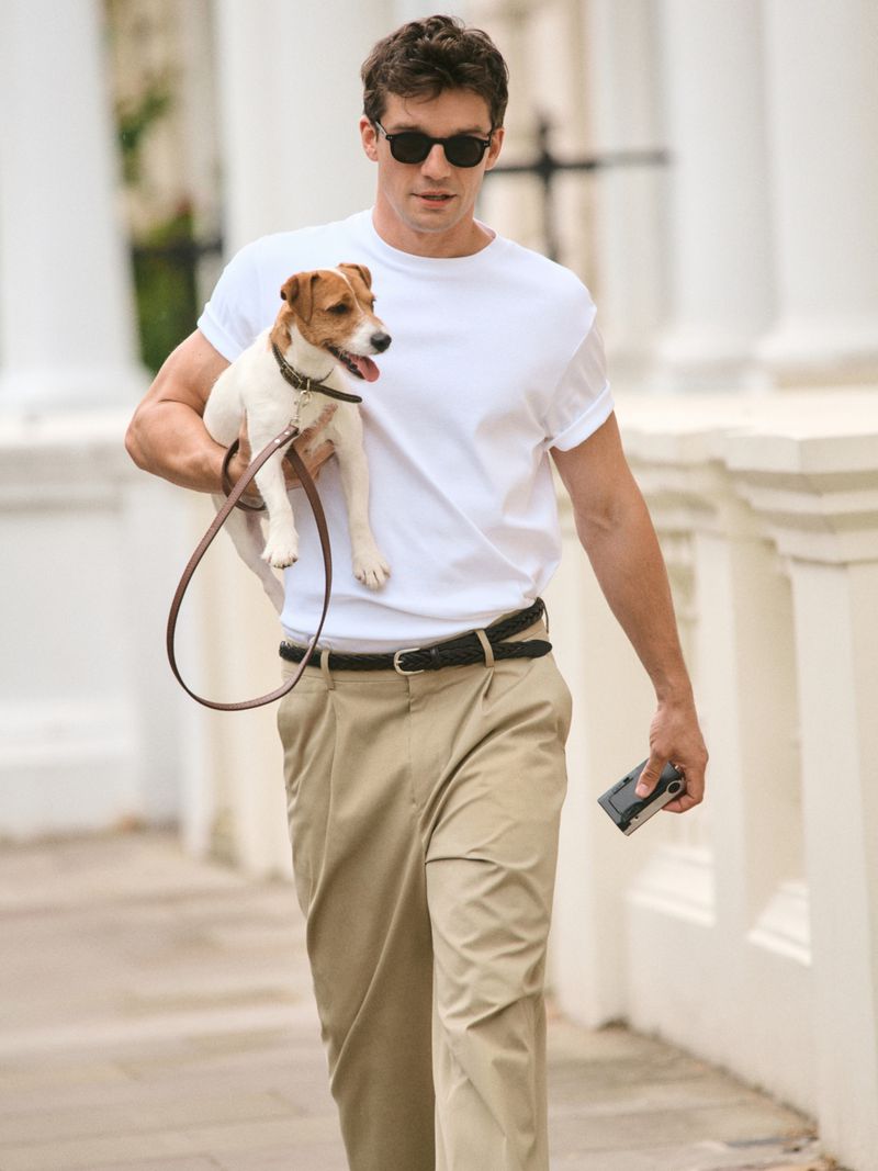 A man in sunglasses walks a Jack Russell Terrier on a leash in a neighborhood with white buildings and trees in the background.