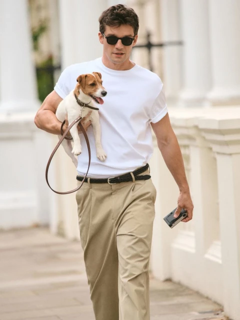 A man in sunglasses walks a Jack Russell Terrier on a leash in a neighborhood with white buildings and trees in the background.