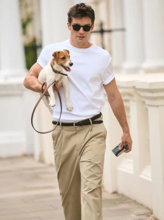 A man in sunglasses walks a Jack Russell Terrier on a leash in a neighborhood with white buildings and trees in the background.