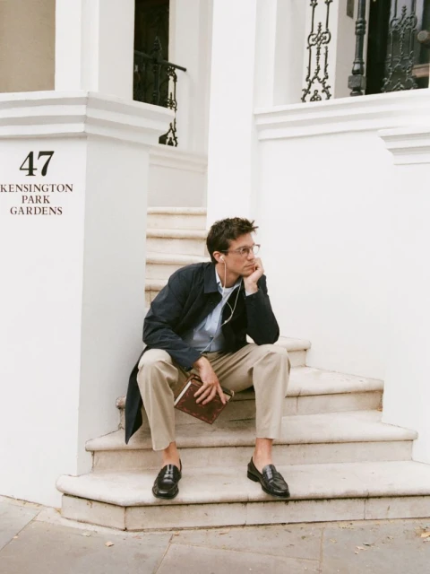 Man in navy coat and glasses sits on white stone steps holding a brown book. He wears a light blue shirt, khaki pants, and black loafers.