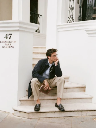 Man in navy coat and glasses sits on white stone steps holding a brown book. He wears a light blue shirt, khaki pants, and black loafers.