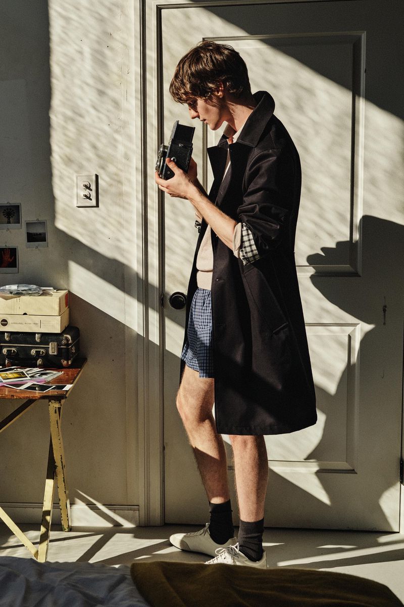 Man in black coat holds vintage camera indoors. Sunlight and shadows play on the white door and wall in the background.