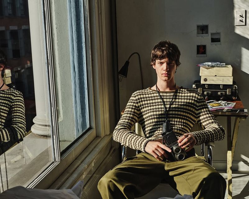 Young adult male with brown hair and a striped shirt poses with a vintage twin-lens camera in a sunlit apartment with a large window.