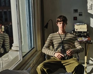 Model Joshua Thompson sitting in a chair holding a vintage camera by a window wearing a striped knit sweater and olive trousers.