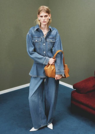 A blonde-haired woman models denim jacket and wide-leg jeans holding a leather bag in a studio with a green wall and blue floor.