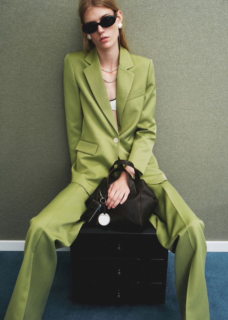 Woman in a green suit and sunglasses sits on a black cabinet, holding a black handbag in a studio setting with a minimalist background.