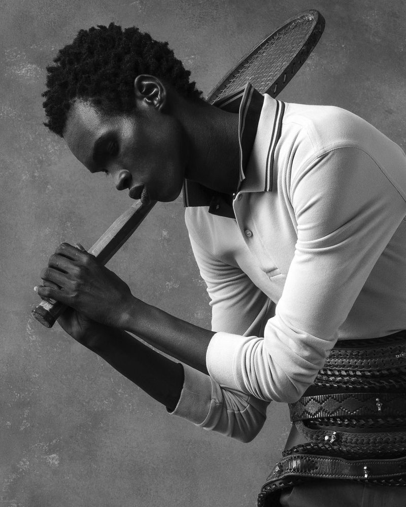 Black and white studio shot of a dark-skinned man in polo shirt holding a tennis racket on his shoulders against a grey background.